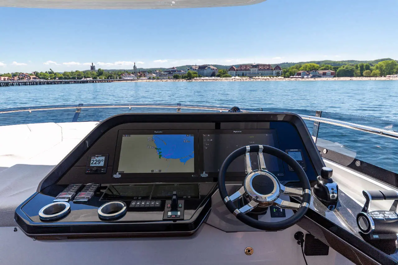 Modern yacht control panel with two screens displaying navigation maps, steering wheel, and scenic waterfront view.