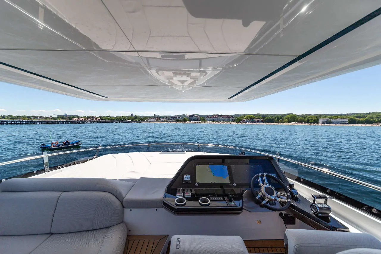 View from a yacht's helm displaying a digital navigation screen and scenic waterfront with a small boat in the foreground.