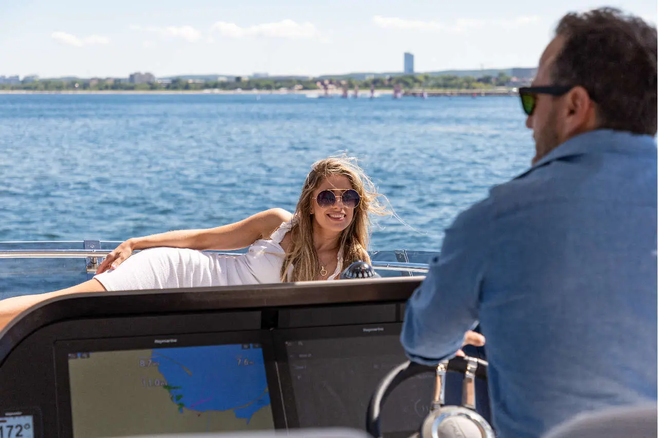 A woman in sunglasses lounges on a boat, smiling at a man steering, with water and a skyline in the background.