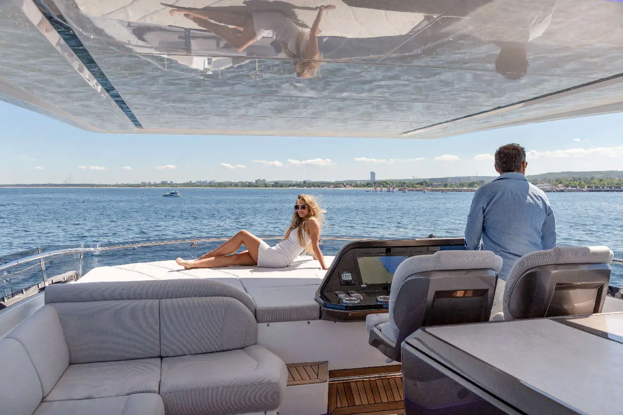 A woman in a white dress relaxes on a yacht, while a man in a blue shirt steers, with a scenic waterfront view.