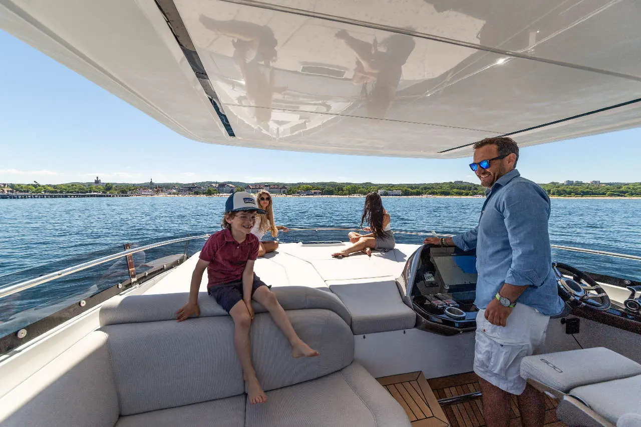 A boy sits on a yacht, smiling, while a man stands at the helm and two women relax on the deck with water in the background.