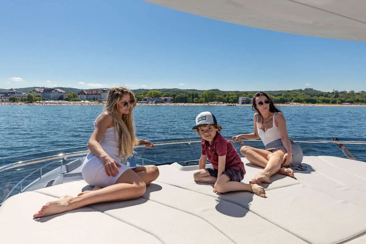 Three people relax on a yacht's deck under a clear blue sky, with a scenic view of the coast and beach in the background.
