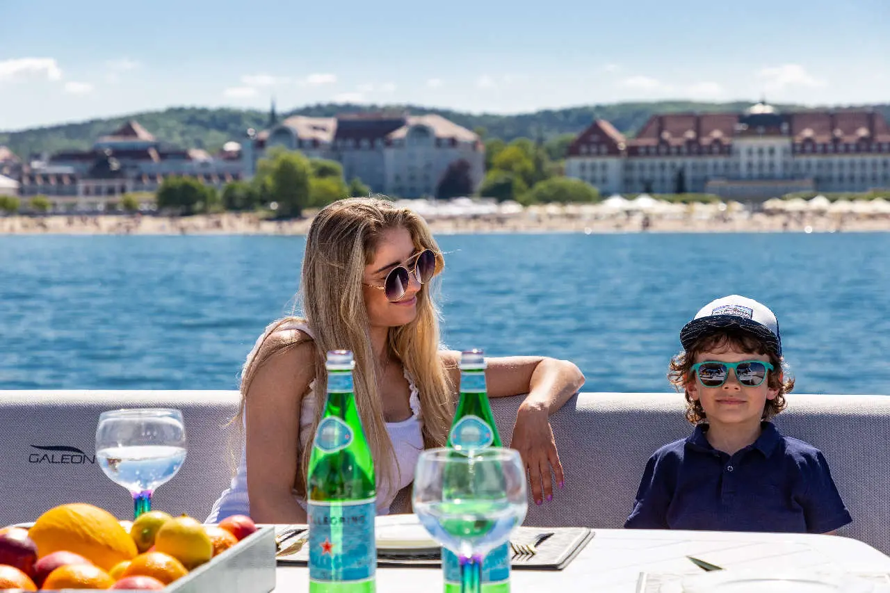 A woman and a boy sit on a yacht, enjoying drinks with a scenic waterfront view and fruit on the table.