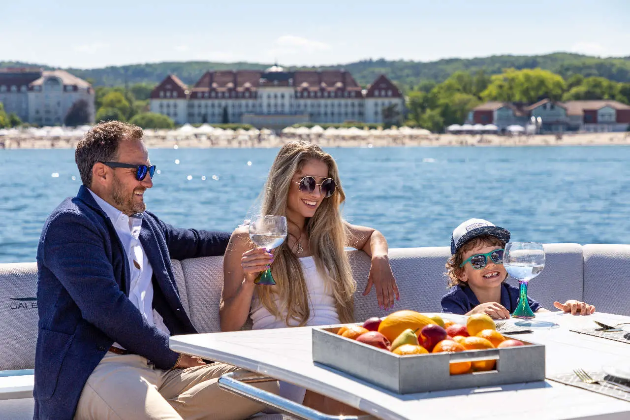 A woman and child enjoy drinks on a boat, with fruit on the table and a scenic hotel view across the water.