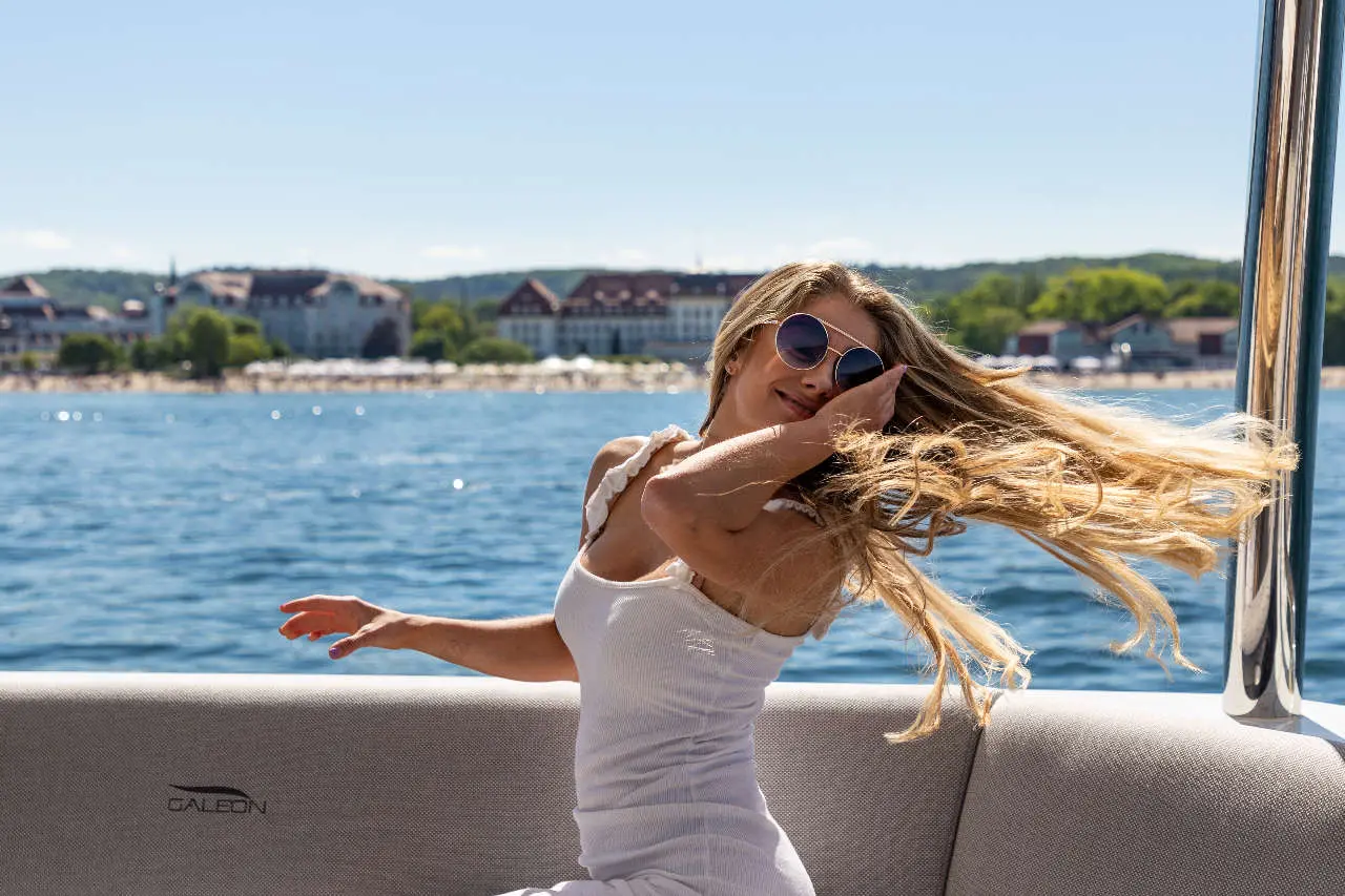 Woman in a white dress, wearing sunglasses, enjoying a sunny day on a boat with sea and coastal buildings in the background.