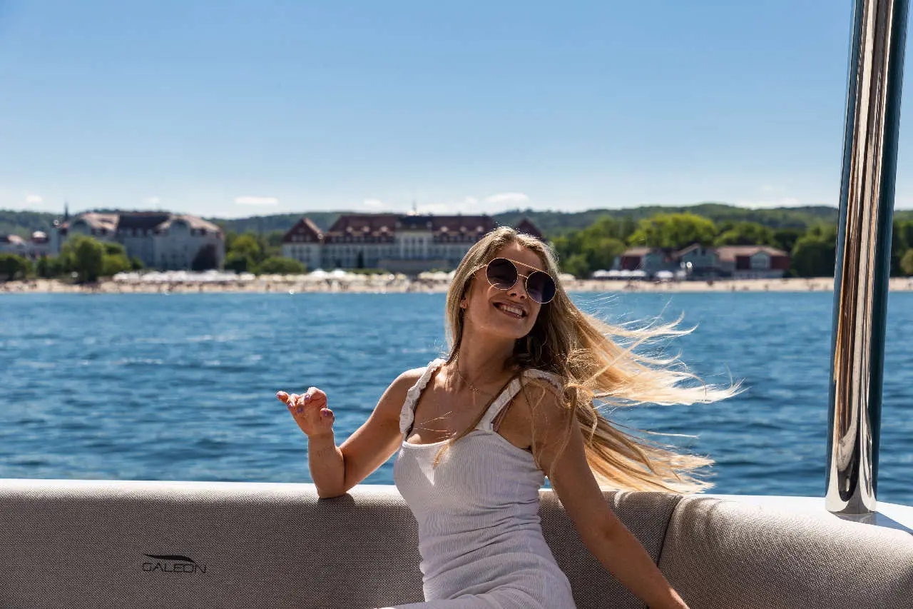 Woman in sunglasses enjoys a sunny day on a boat, with waterfront resorts and water in the background.