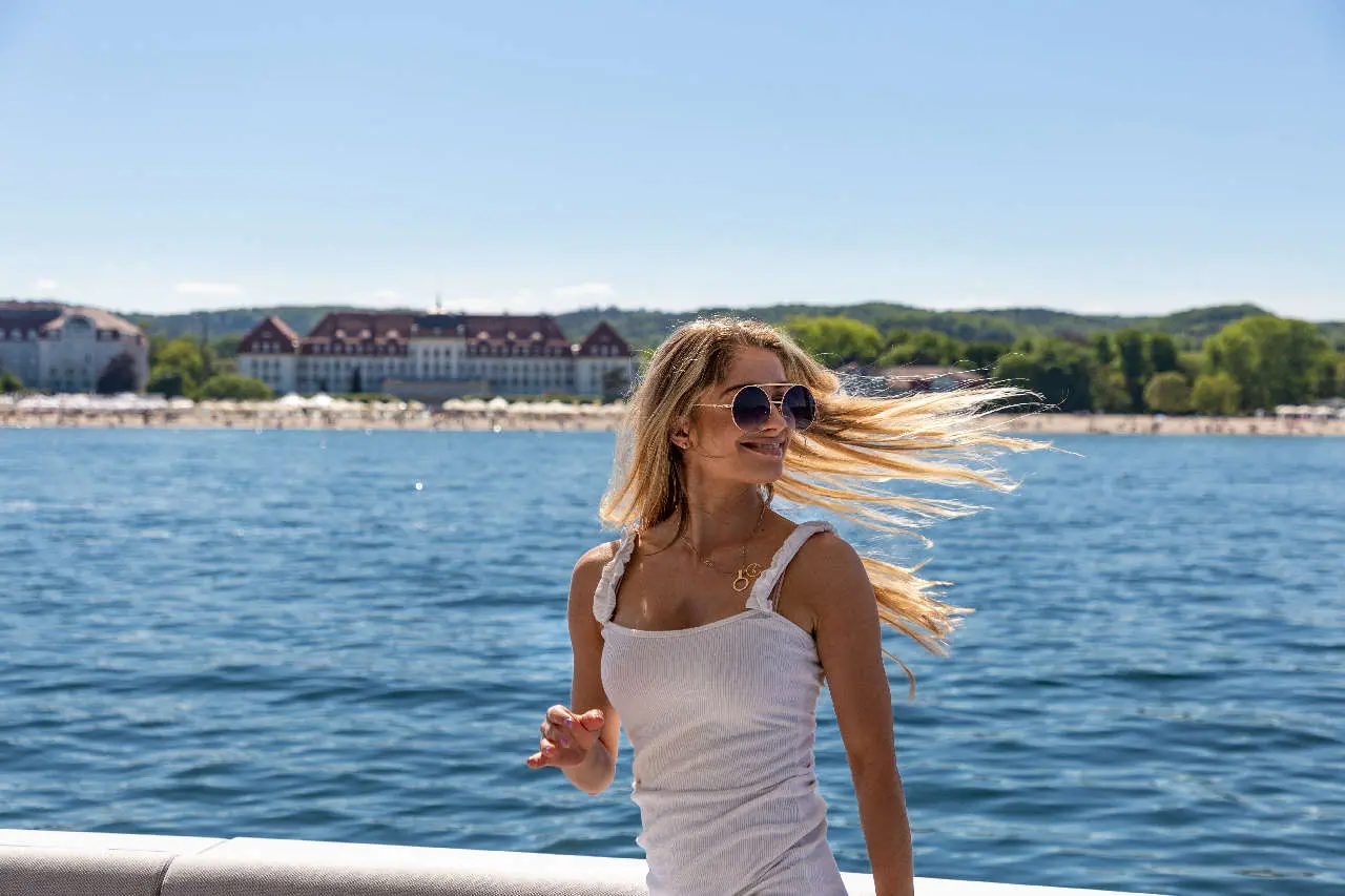 Woman in sunglasses and white dress smiles by the waterfront, with a beach and hotel in the background under clear blue skies.