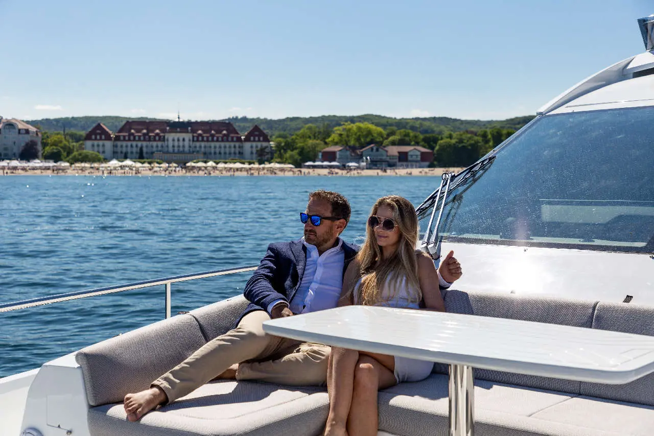 A couple relaxes on a yacht, overlooking a beach and historic hotel, enjoying a sunny day by the water.