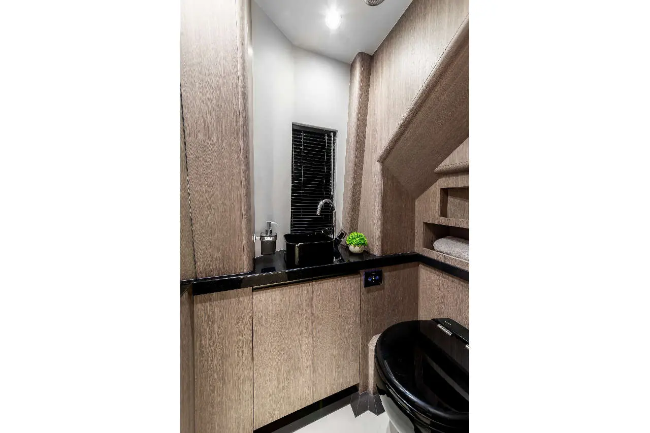 Modern bathroom featuring wooden cabinetry, black sink, toilet, and a decorative plant, with sunlight filtering through blinds.
