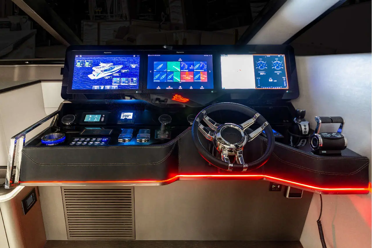 A sleek boat console features dual screens displaying navigation and controls, a stainless-steel steering wheel, and illuminated panels.
