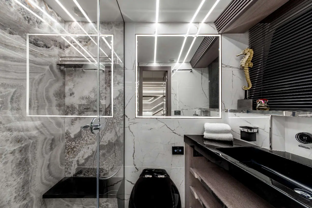 Modern bathroom featuring grey marble walls, a glass shower, and sleek black fixtures. Lit mirrors and decorative elements enhance elegance.