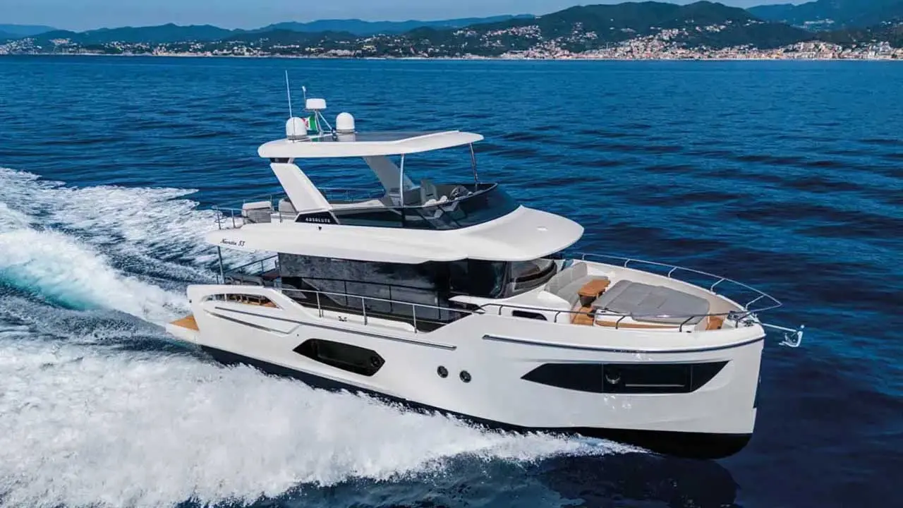 White luxury motor yacht cruising at speed, leaving a foamy wake with a mountainous coastal town in the background.