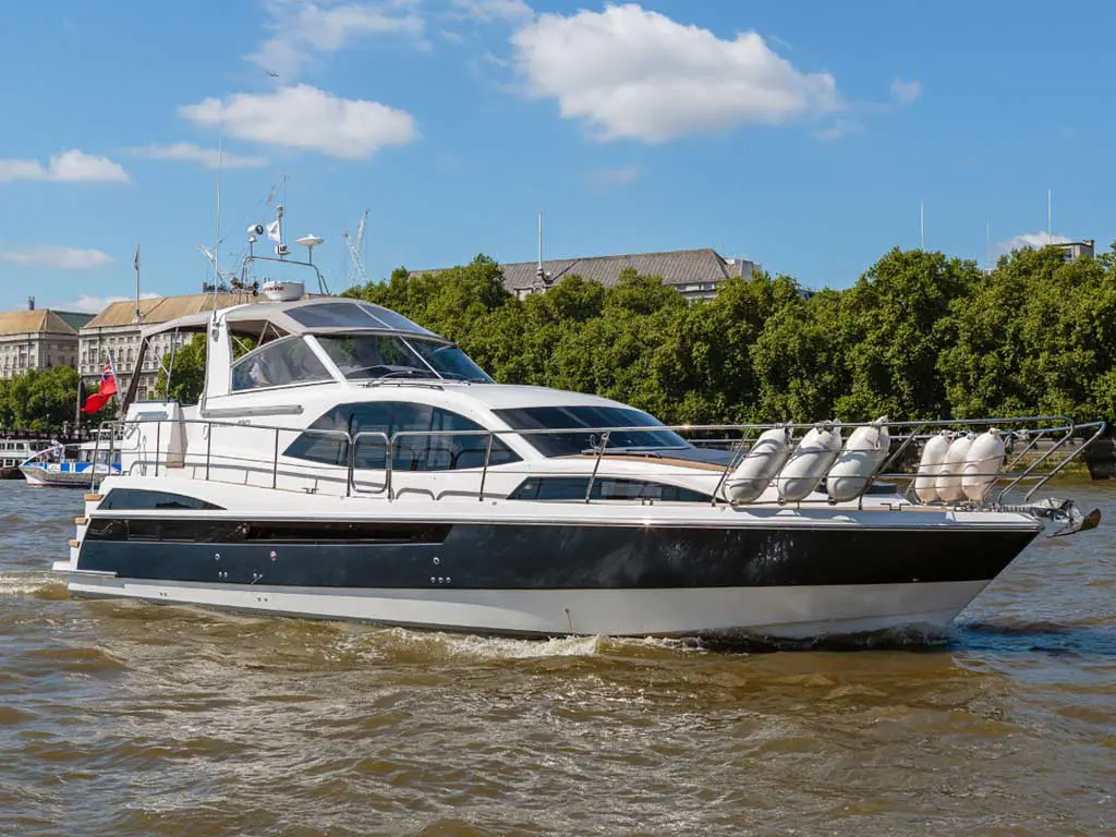 A sleek motor yacht navigates a river, with a clear blue sky and lush greenery lining the banks.