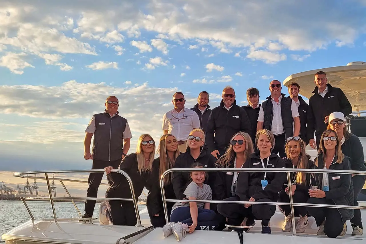 Group photo of 15 people on a boat during sunset, all wearing matching dark jackets, smiling and enjoying the moment.