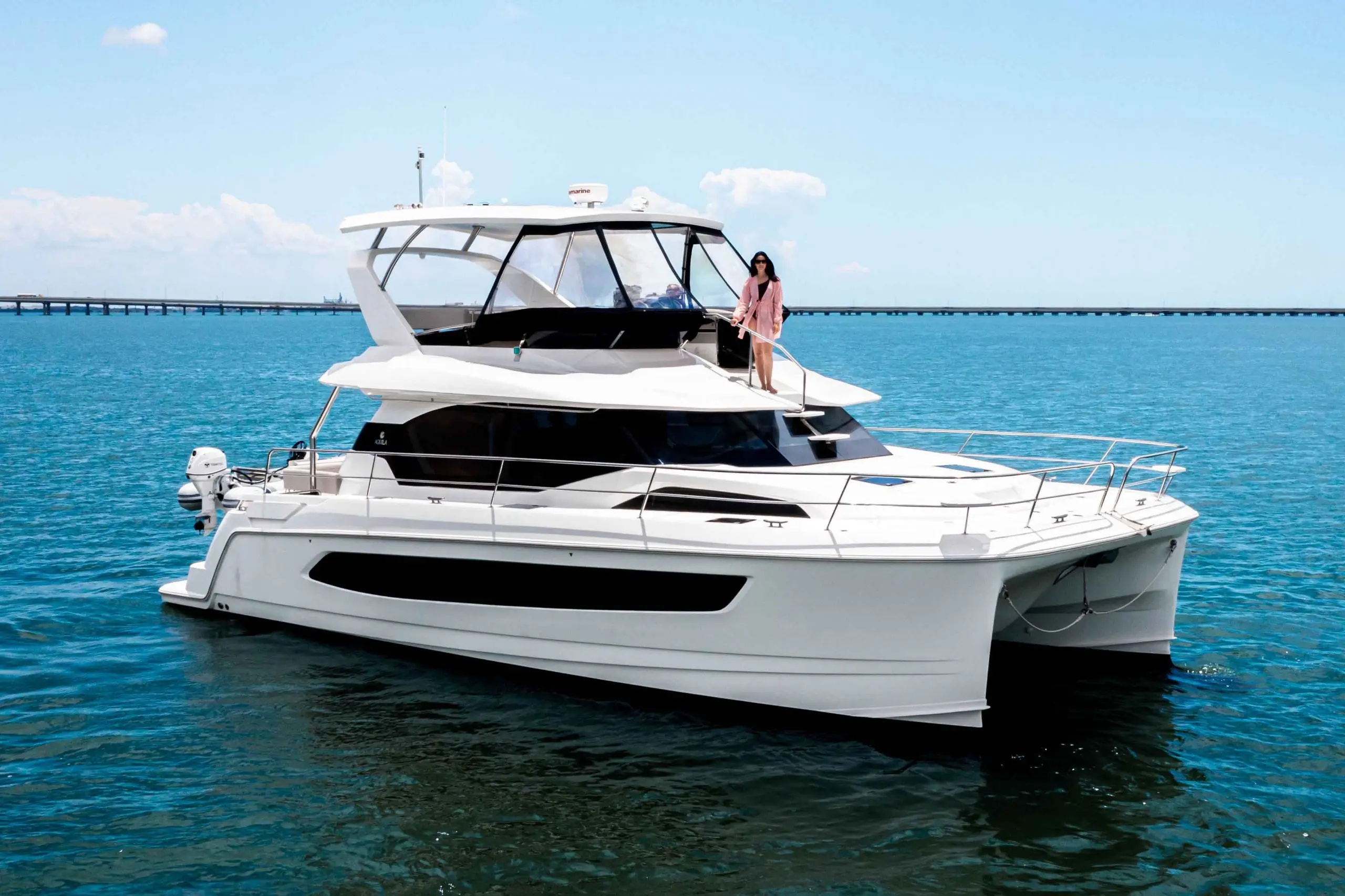 A stylish white catamaran with a woman standing on the bow, moored in calm turquoise waters under a blue sky.