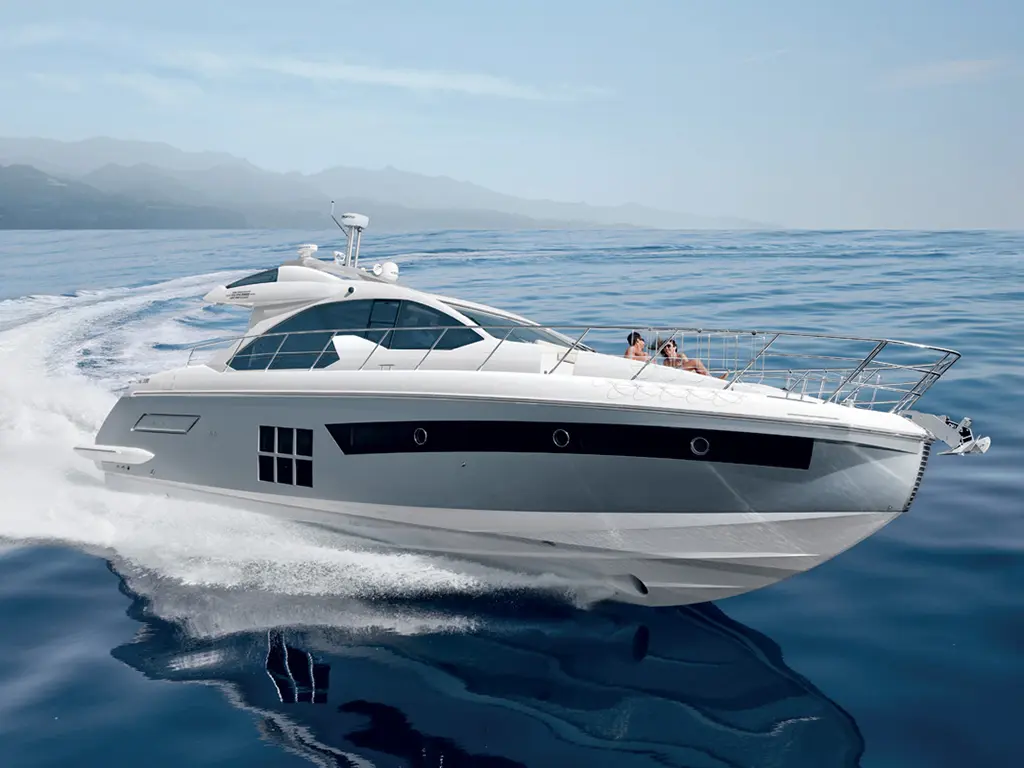 A sleek white yacht speeds across calm blue waters, with two people relaxing on the bow under a clear sky.