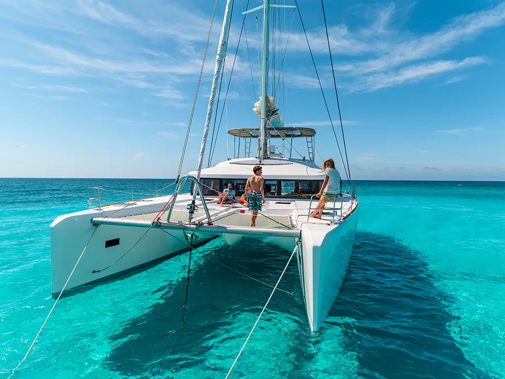A catamaran anchored in clear turquoise water with three people enjoying the sun on deck under a blue sky.