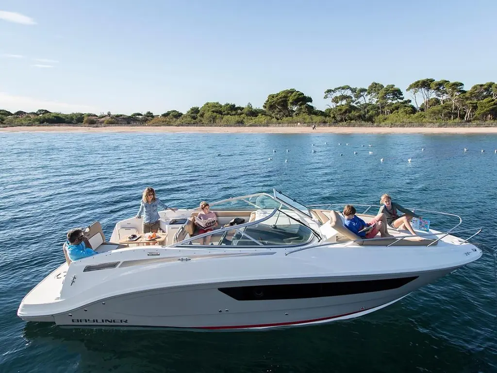 A group of four people relax on a Bayliner boat in serene waters near a sandy beach, enjoying the sun and each other's company.