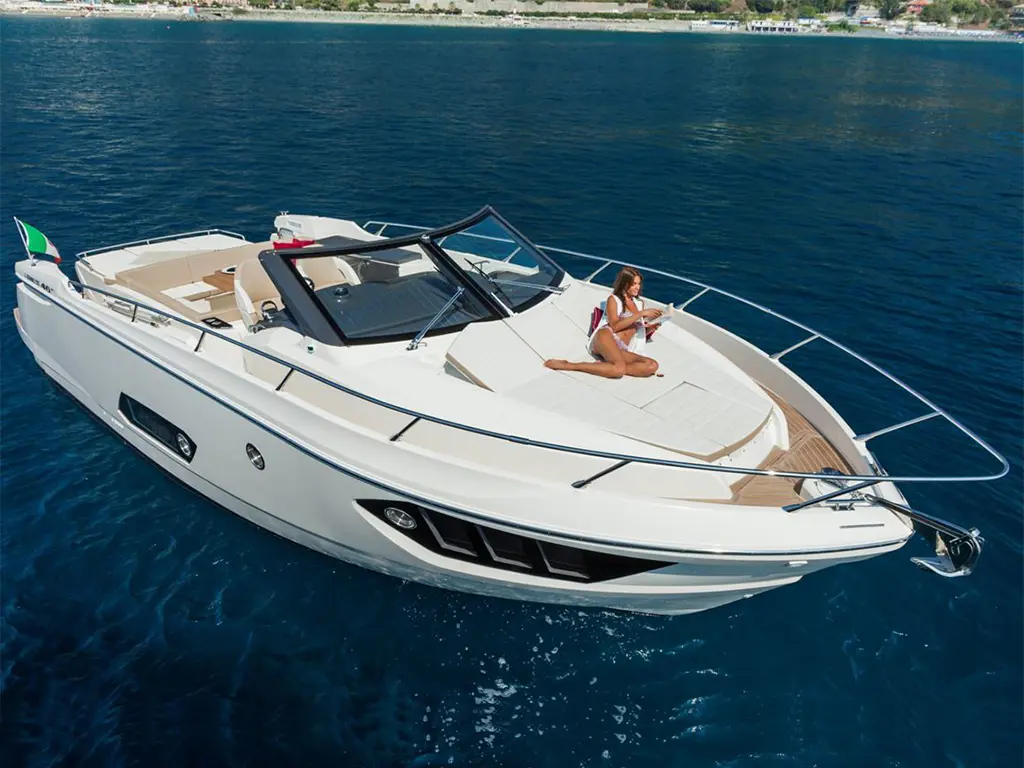 A sleek white motorboat glides on blue water, with a woman in a bikini relaxing on the deck, reading a magazine.