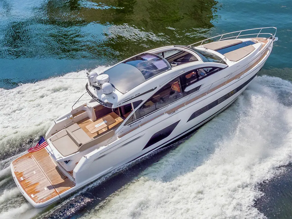 A luxury yacht cruises over waves, displaying a polished white exterior and wooden deck, with an American flag at the stern.