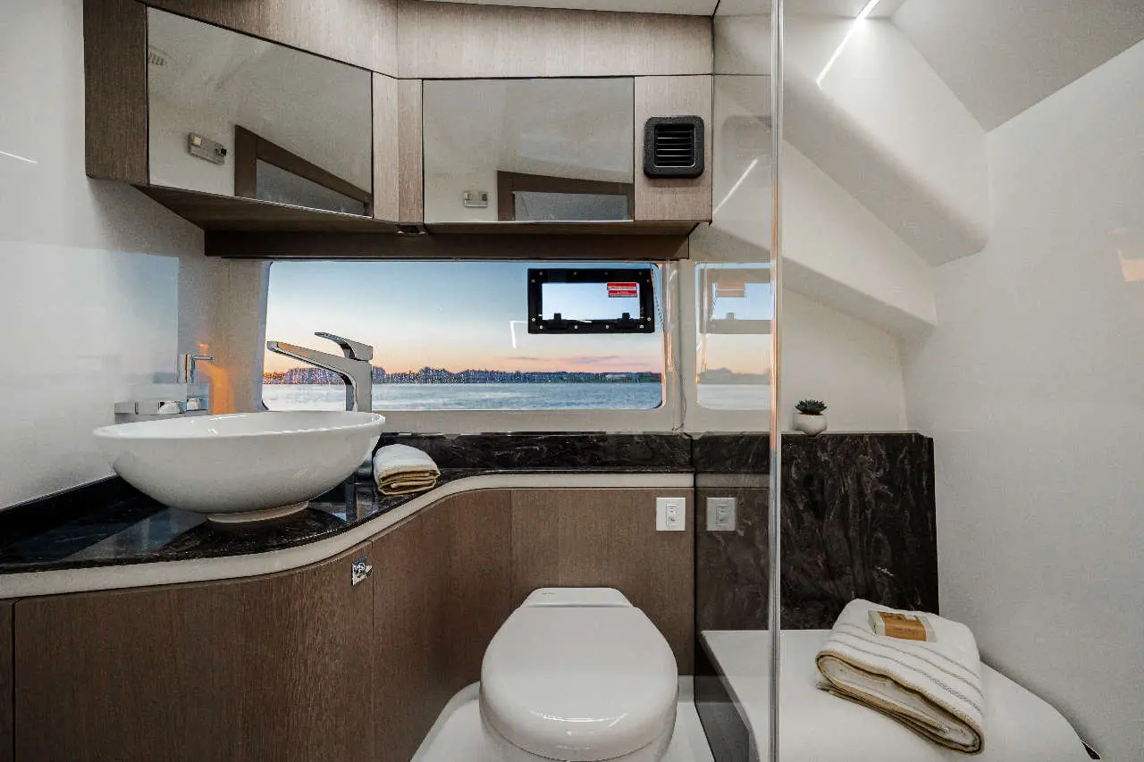 Modern boat bathroom featuring a circular sink, toilet, and scenic window view of the water and sunset. Neat decor elements visible.