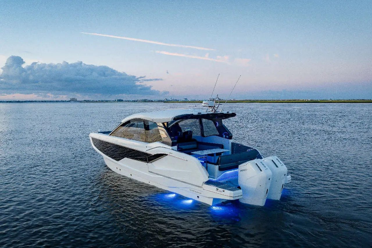 Sleek motorboat with blue under-lighting on water at dusk, featuring a spacious cockpit and city skyline in the background.