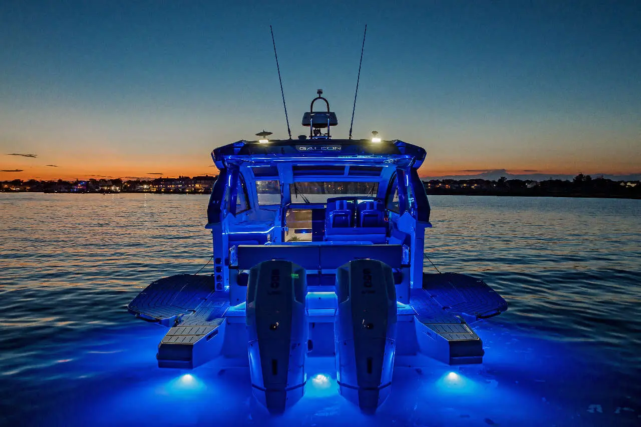 A lit-up boat with twin engines at dusk, facing a calm waterway and a vibrant sunset sky in the background.