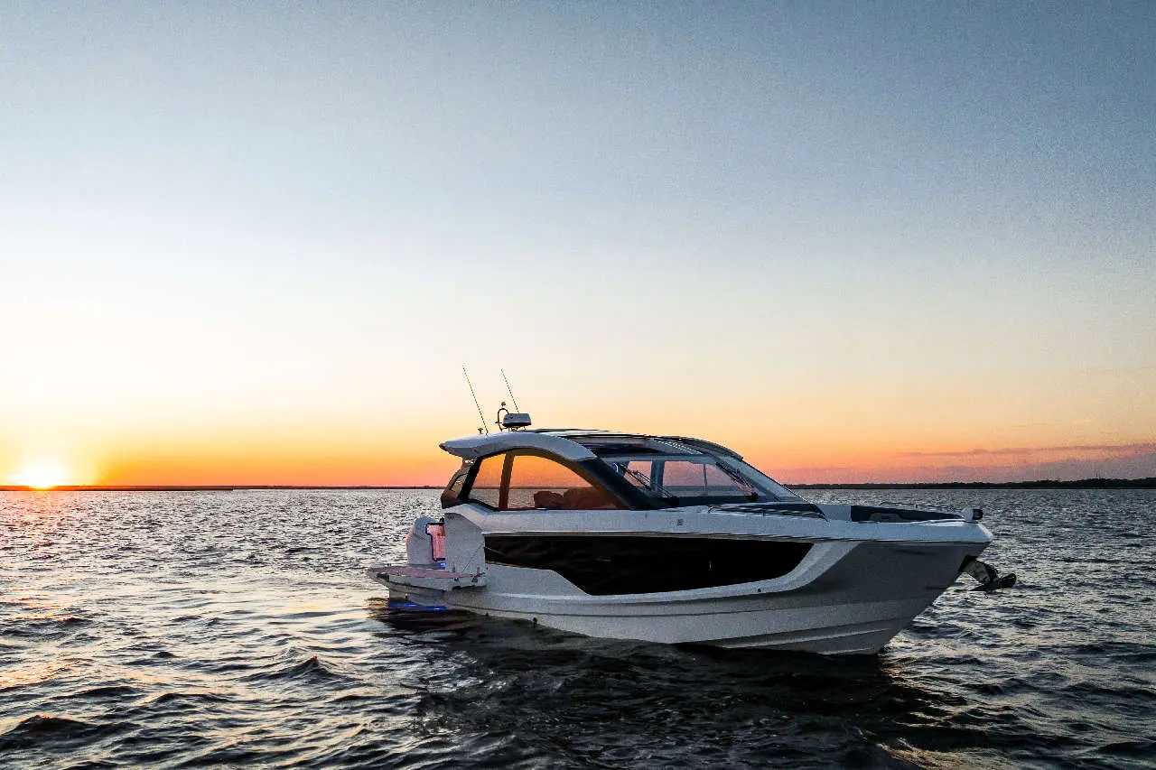 A sleek white motorboat navigates calm waters at sunset, with a warm orange sky and distant horizon visible.