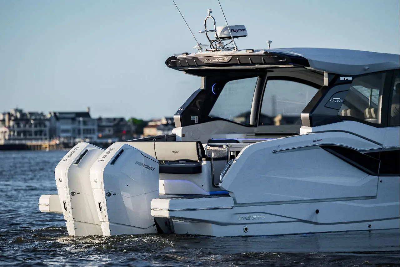 A luxury white boat with twin Mercury outboard motors navigating calm waters, with a waterfront neighbourhood in the background.
