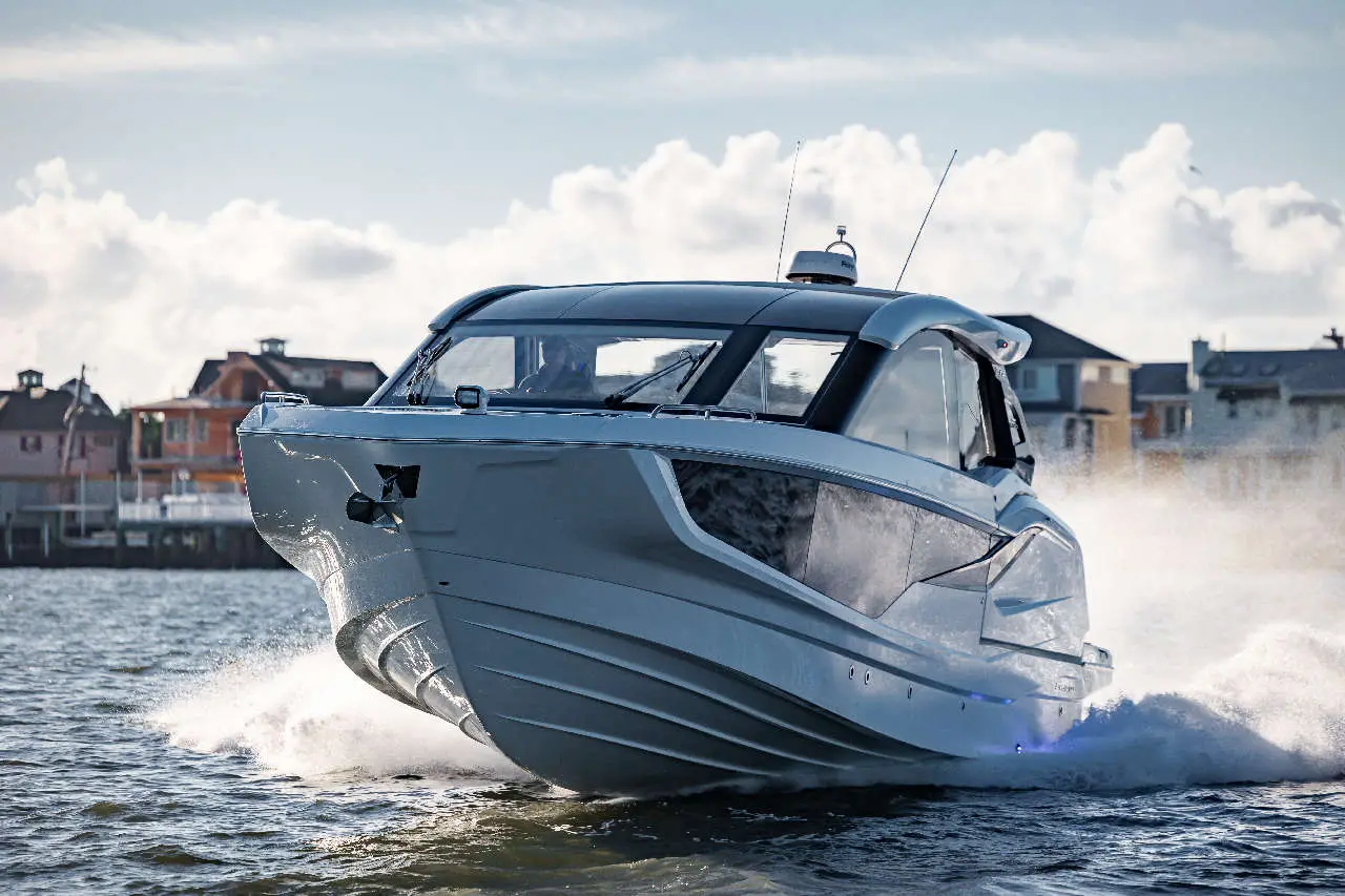 A sleek powerboat speeds across the water, creating splashes, with homes visible along the shoreline in the background.