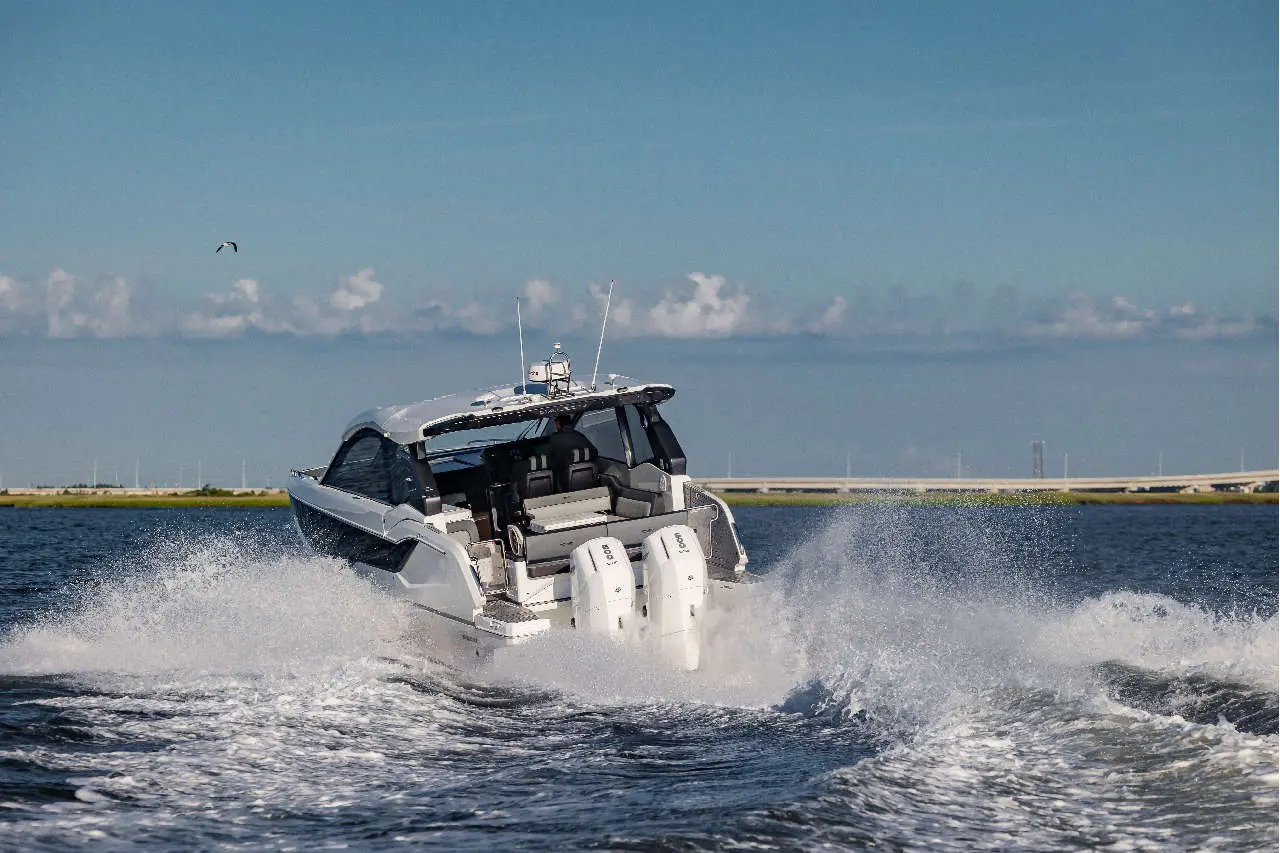A powerboat speeds across the water, creating a spray, with a clear sky and distant bridge in the background.
