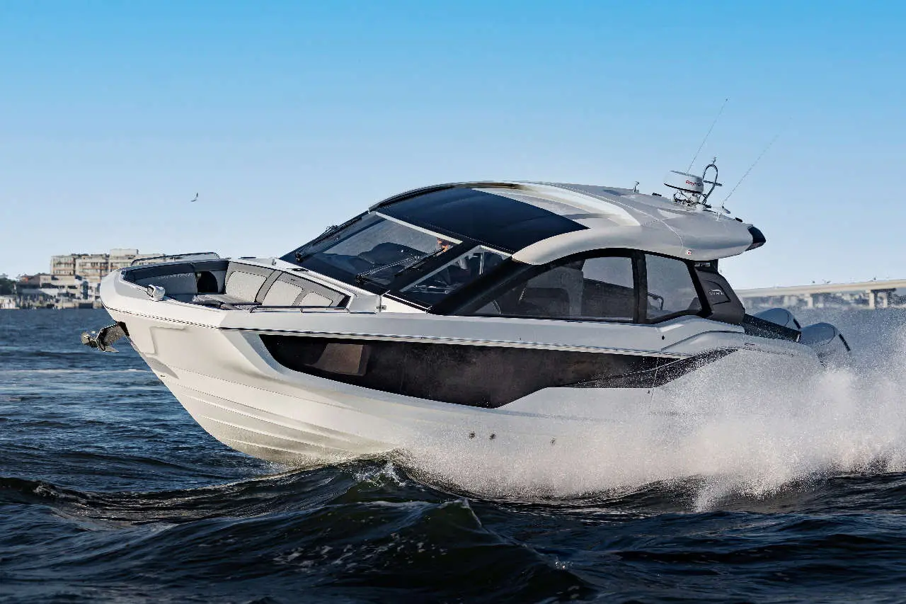 A sleek motorboat skims across the water, creating splashes, with a clear sky and buildings in the background.