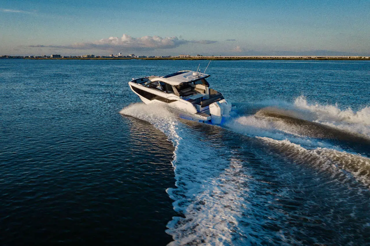 A sleek white motorboat speeds across calm waters, creating a spray against a clear sky and distant urban skyline.