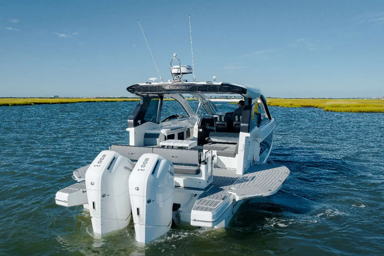 A modern powerboat with twin outboard motors glides through calm water, with a person seated at the helm and lush greenery in the background.