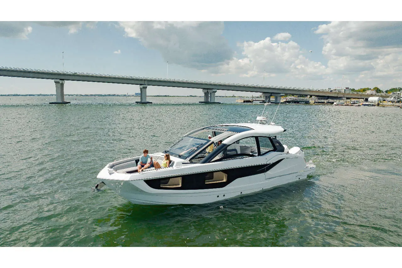 A sleek white yacht glides through calm waters near a bridge, with two people relaxing onboard in sunny weather.
