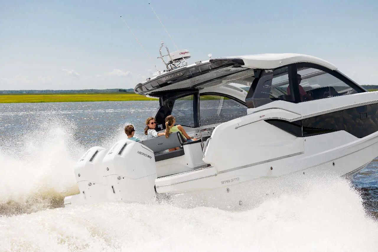 A sleek white motorboat with children enjoying a ride, creating splashes in a scenic waterway. Mercury engines visible.