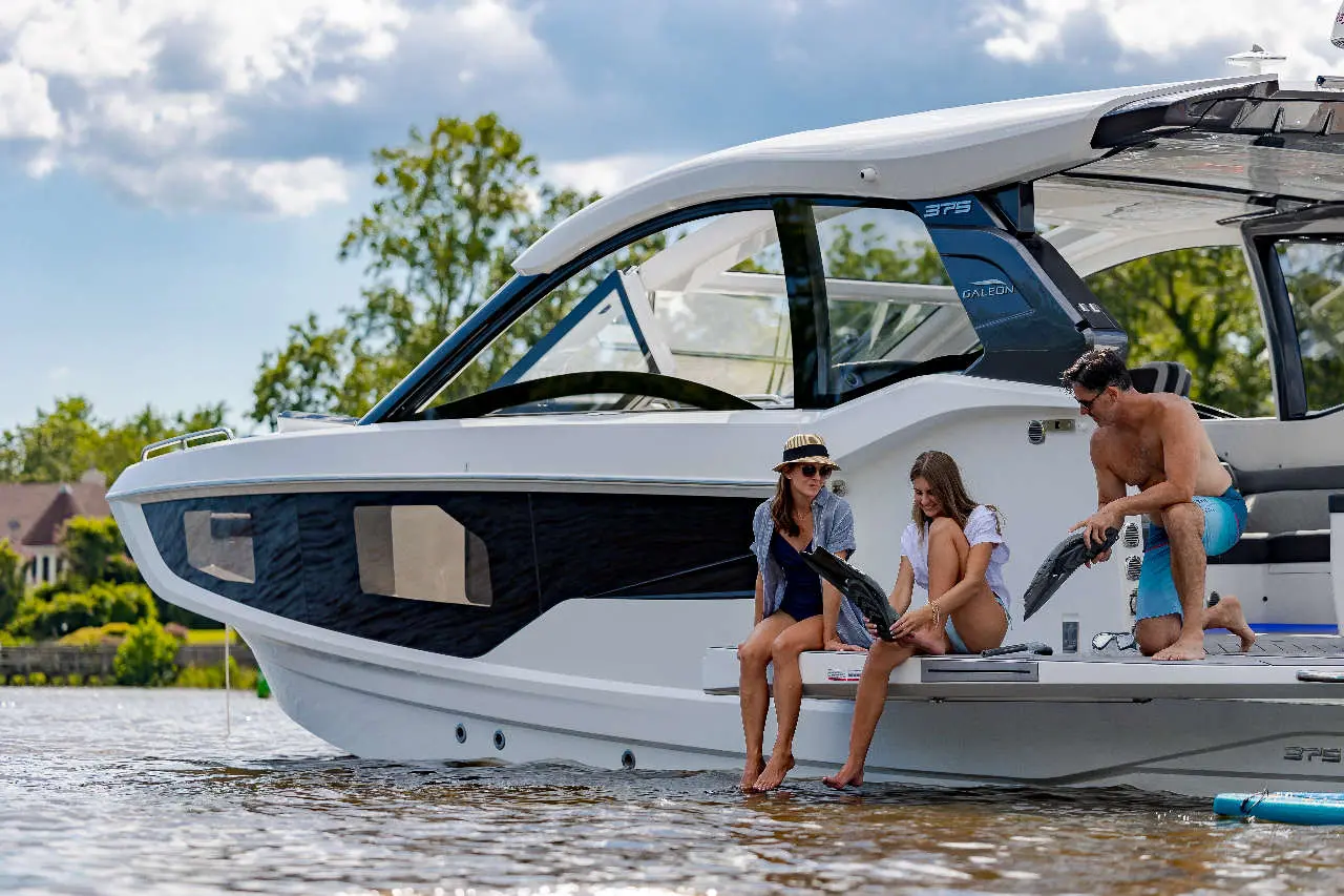 Three people relax on the edge of a sleek white motorboat, preparing water sports gear on a sunny day by the river.