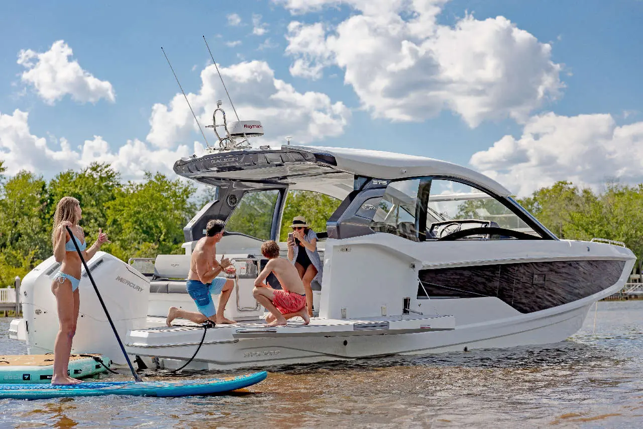 A family enjoys summer by a sleek motorboat, with a girl paddleboarding and two children engaging with adults on board.