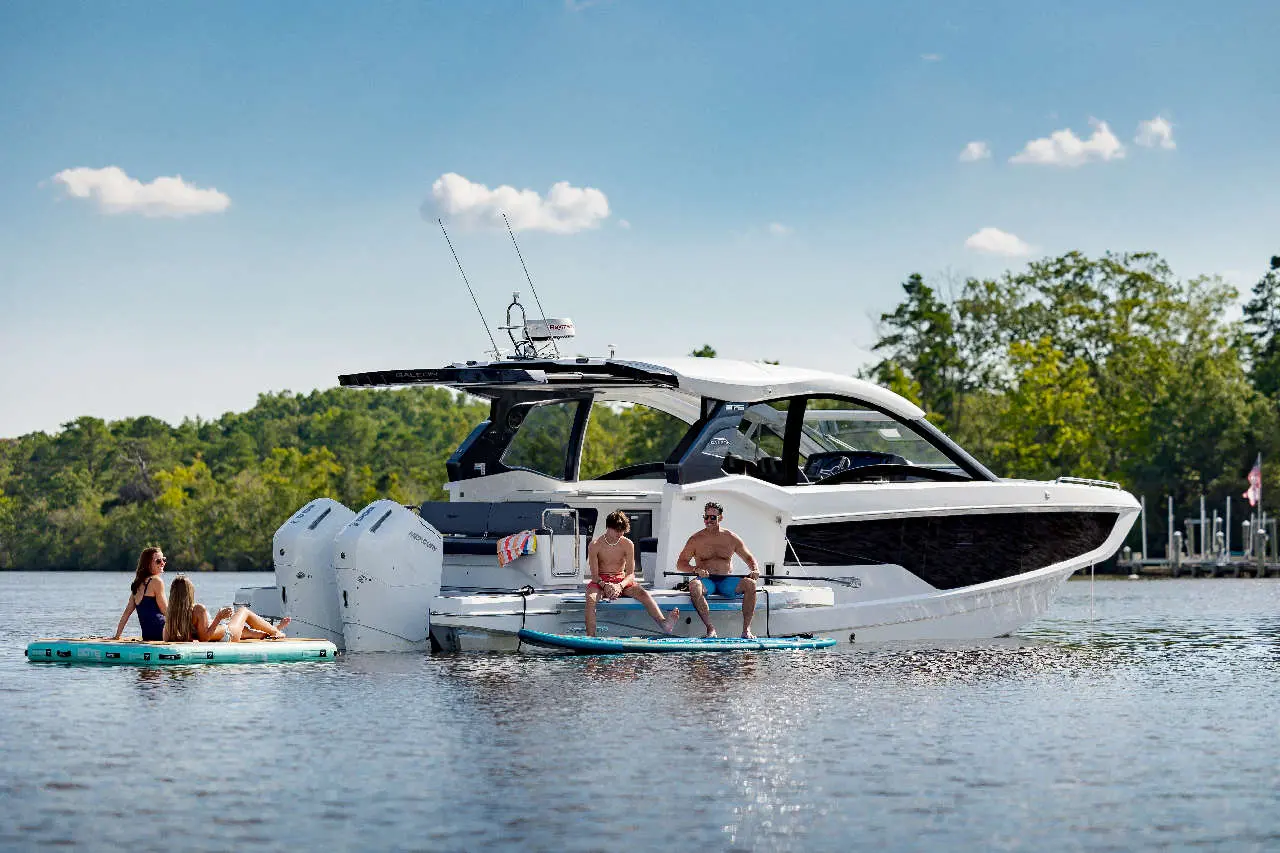 A luxury boat floats on calm water with two women on a paddleboard and two men seated nearby, enjoying the sun.