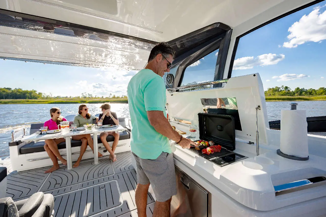 Man grilling skewers on a boat while three friends enjoy a meal at a table, with a scenic river view and clear skies.