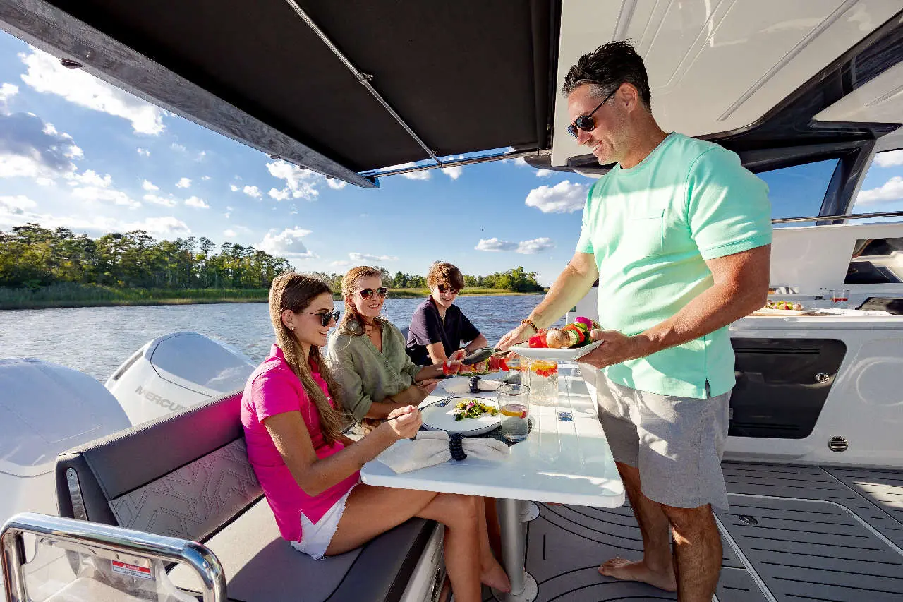 A man serves food to three friends dining on a boat, enjoying a sunny day by the water with scenic views.