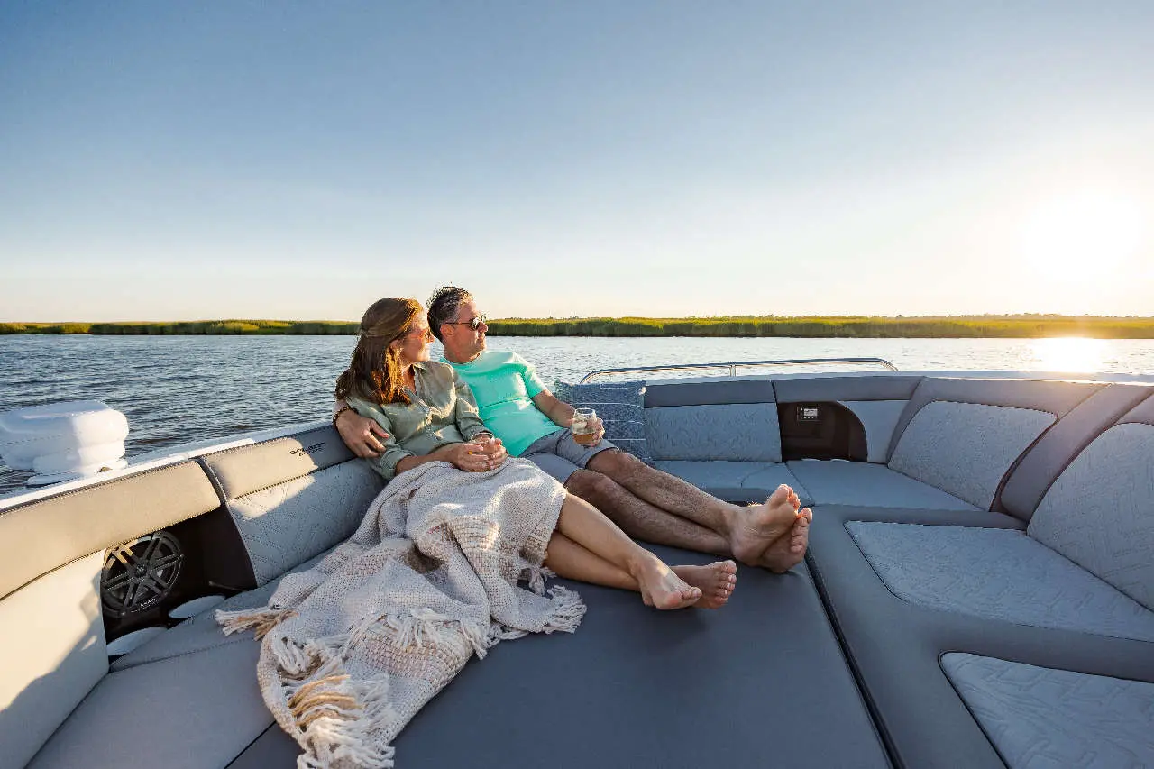 A couple relaxes on a boat, wrapped in a blanket, enjoying drinks by the water at sunset.