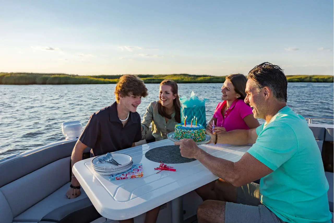Group celebrating a birthday on a boat, with a cake and gifts, enjoying the scenic waterfront view.