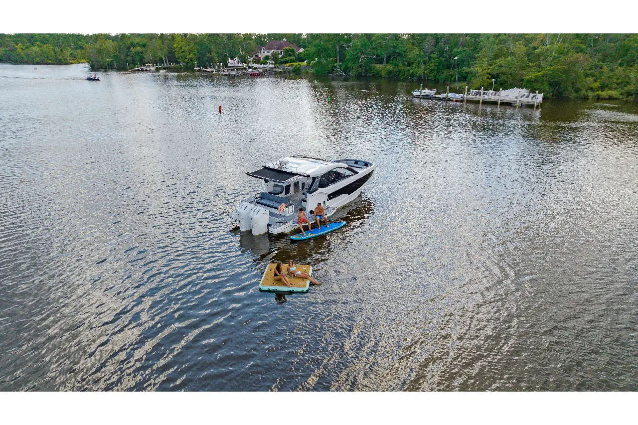 Two people on a paddleboard and a floating mat near a luxury boat on calm waters, surrounded by trees and docks.