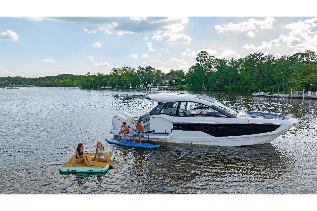 Two adults and two teenagers relax on floating mats near a luxury boat on a calm river surrounded by greenery.