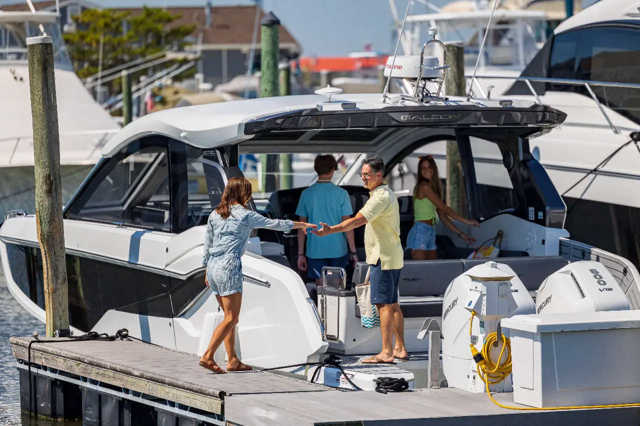A woman in denim approaches a man on a yacht, while two others stand nearby at the dock.
