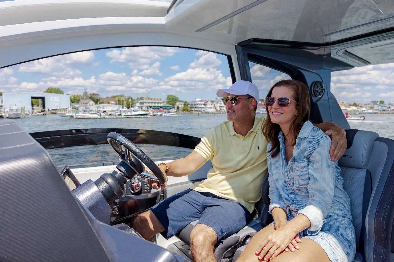 A man and woman enjoy a boat ride, smiling as they sit in the cockpit with scenic waterfront in the background.