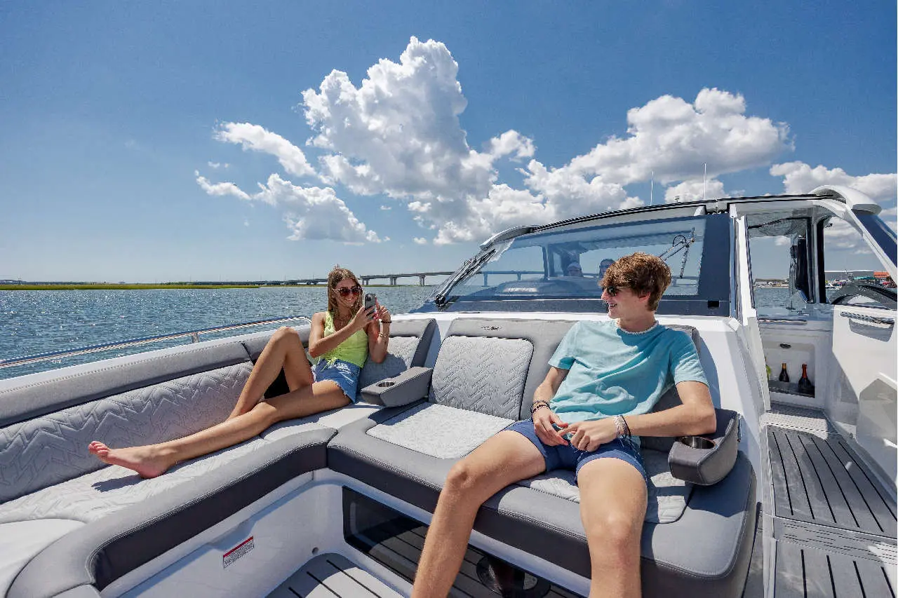 Young woman in a green top sits on a boat, using her phone, while a young man in a blue shirt relaxes nearby.