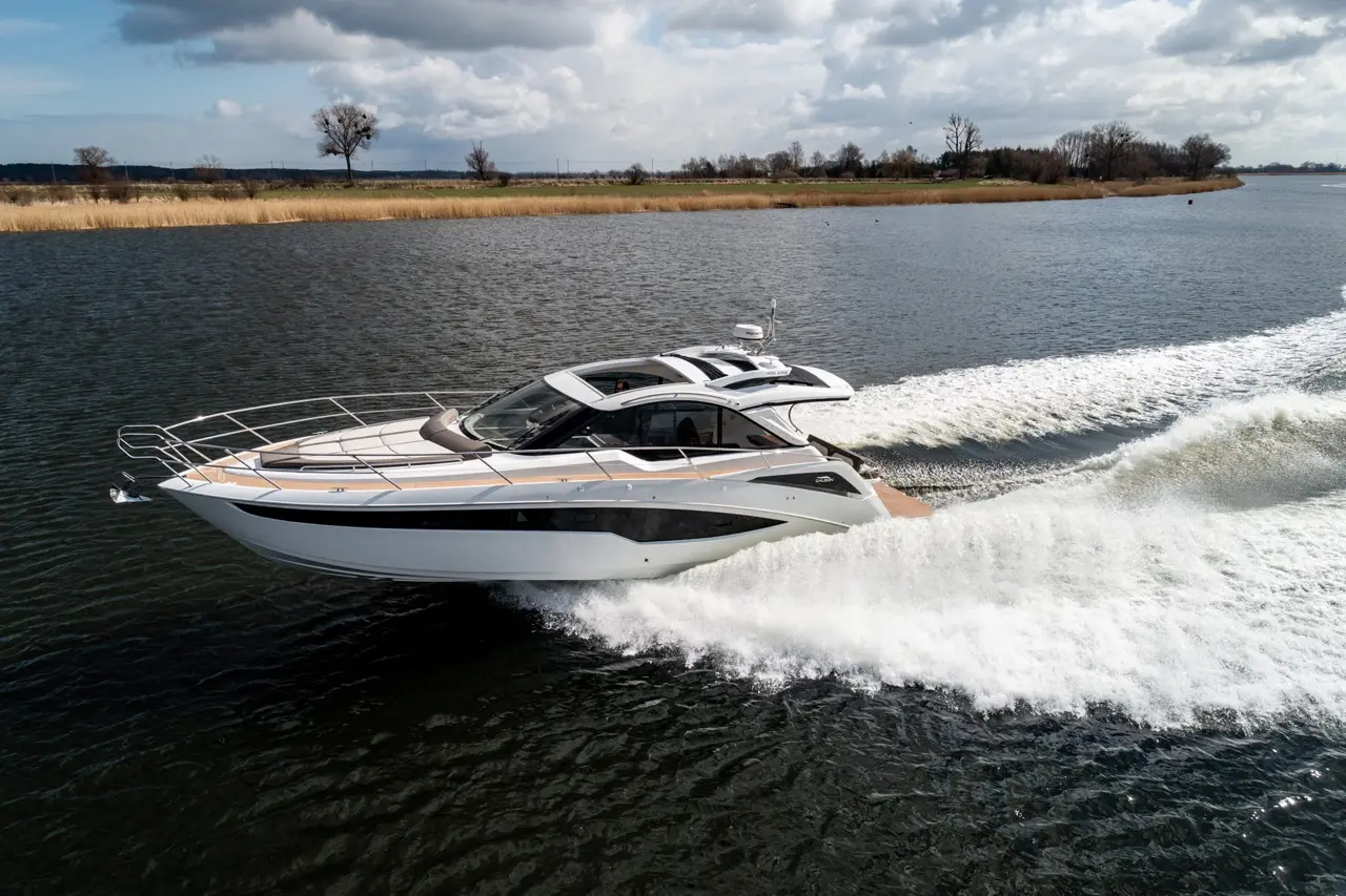 A sleek white motorboat glides across a calm waterway, creating a spray of white foam against a cloudy sky backdrop.