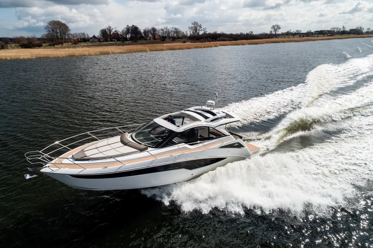 A sleek motorboat speeds across a calm river, leaving white water trails behind, with trees and shore in the background.